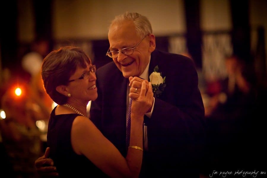 An elderly couple, wearing formal attire, shares a joyful dance at a dimly lit event. The man holds the woman’s hand and smiles warmly, wearing glasses and a suit with a white rose boutonniere. She wears a sleeveless dress and pearls.