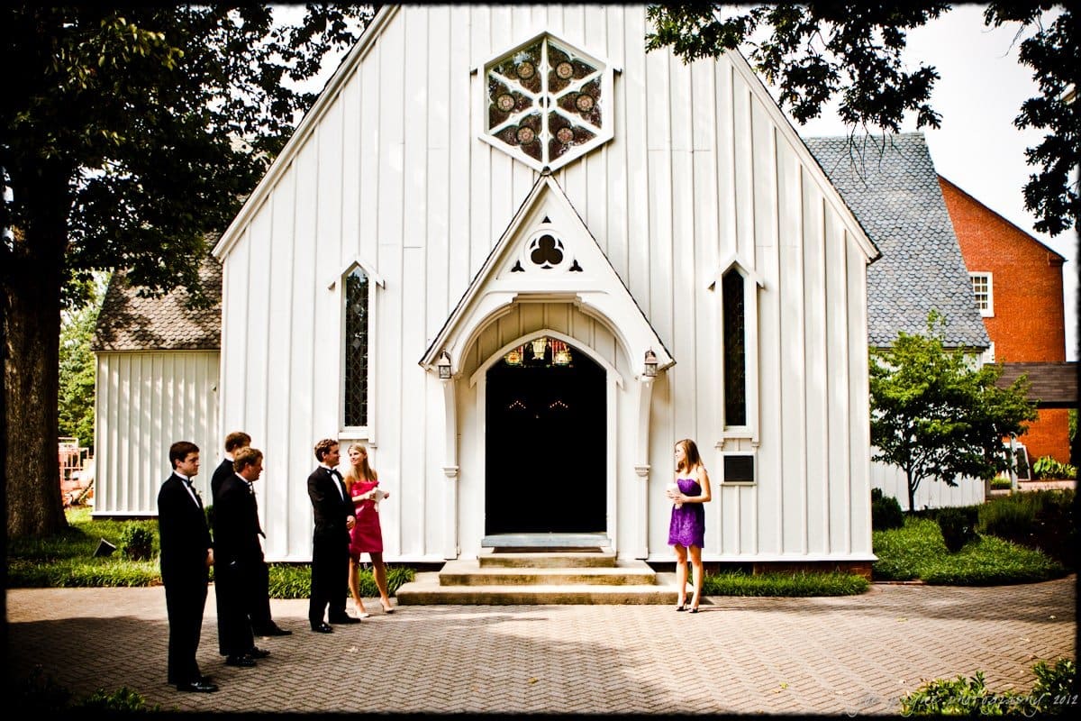 A group of well-dressed individuals stand outside a small white chapel with a triangular archway. The men are in tuxedos, while the woman wears a purple dress. A colorful stained glass window is seen above the entrance. Trees and buildings surround the chapel.