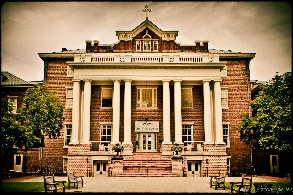 A large, historic brick building with classical columns and a cross on top. The entrance has steps leading up to it, flanked by trees and a sign reading Welcome to Saint Marys School. Benches are in the foreground.