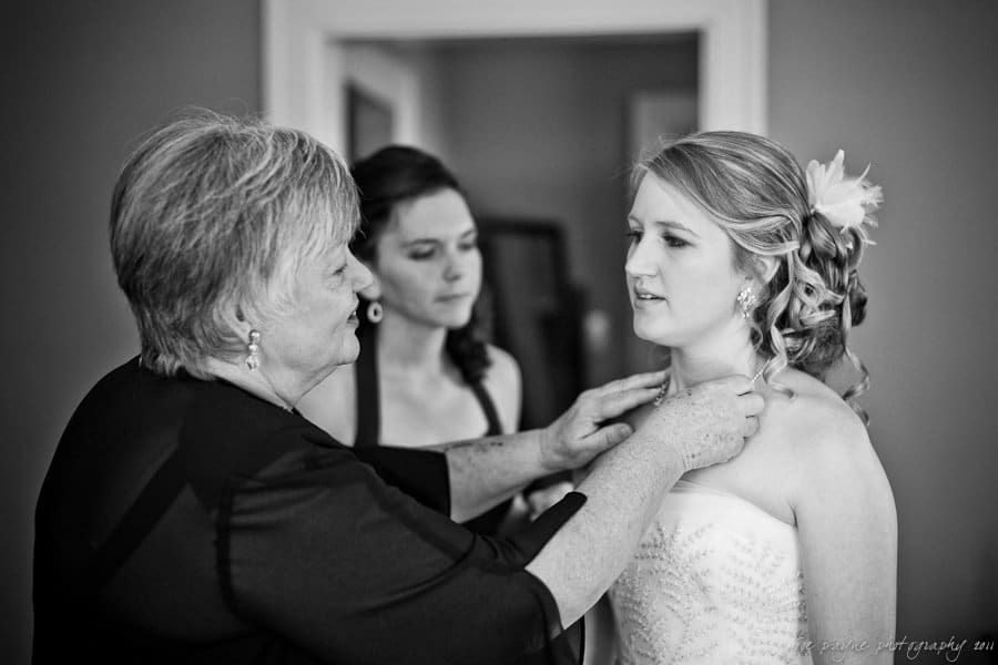 A bride in a strapless dress stands still as an older woman helps adjust her necklace. Another woman looks on in the background. The scene is set indoors with a softly lit, intimate atmosphere.
