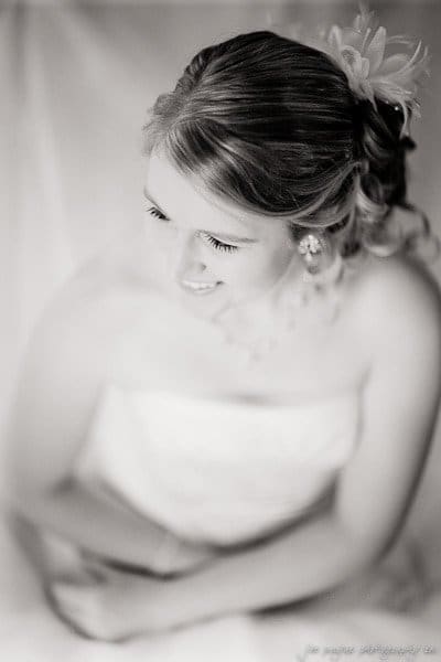 A black and white photo of a bride smiling and looking downward. Her hair is styled with curls and a flower accessory. She wears a white strapless dress and jewelry. The image has a soft, dreamy focus.