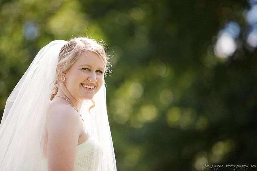 A smiling bride with a long veil stands outdoors against a blurred background of greenery, bathed in sunlight.