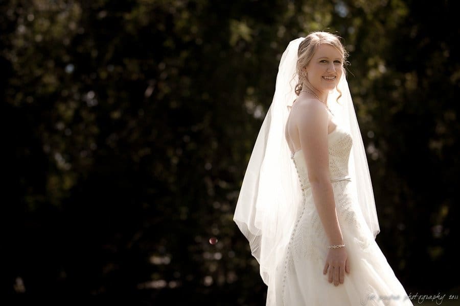 A bride in a strapless white wedding dress and veil smiles while standing outdoors with trees in the background.