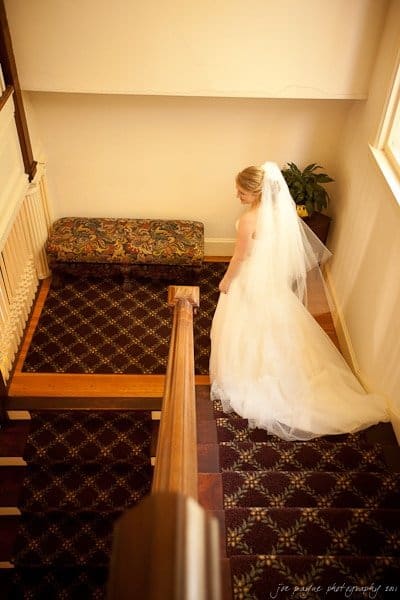 A bride in a white dress and veil descends a carpeted wooden staircase. She is in a softly lit, warm-toned room, walking toward a corner with a small bench and plant. The scene is serene and intimate.