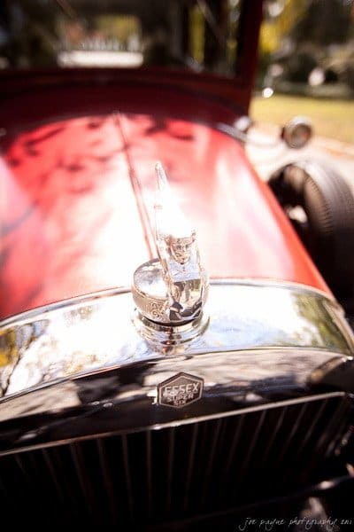 Close-up of the front of a vintage red Essex car. The focus is on the hood ornament and the Essex emblem above the grille. The car is parked outdoors, with sunlight creating reflections on the chrome details.