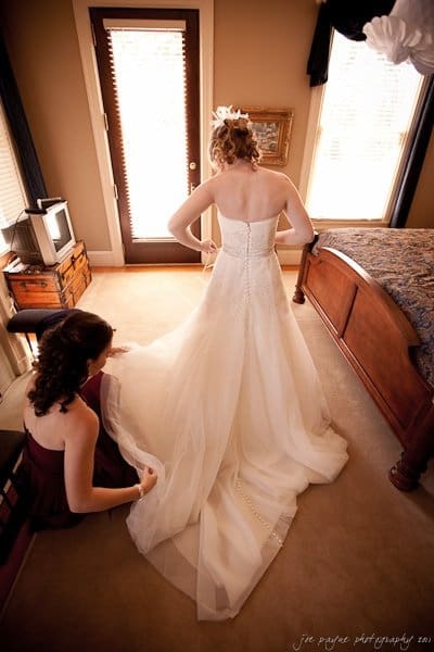 A bride in a white gown stands in a warmly lit room, being assisted by a woman in a burgundy dress who adjusts the gowns train. Sunlight streams through a door in the background, creating a serene atmosphere.
