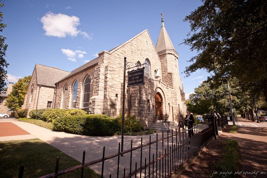 Stone church with a tall steeple and arched windows sits beside a tree-lined street. A black sign in front reads The Cathedral of the Sacred Heart. A few people are near the entrance under a clear blue sky.
