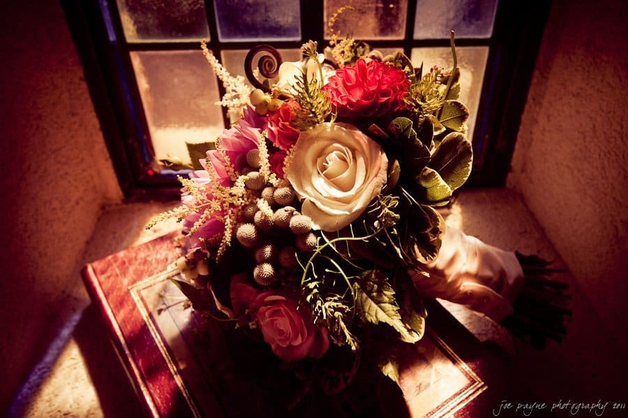 A vibrant bouquet of roses and mixed flowers with greenery is placed near a window, casting a warm light and shadow. The arrangement rests on a vintage book, creating a rustic, romantic atmosphere.