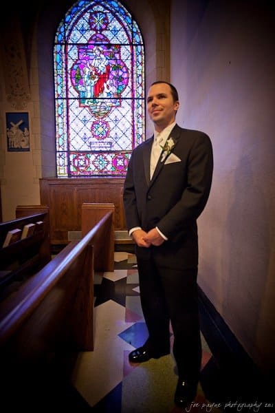 A man in a black suit stands in a church near wooden pews. Behind him is a colorful stained glass window depicting a religious scene. The lighting casts a warm glow on the floors geometric tile pattern.