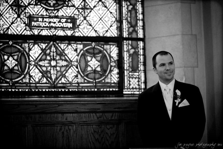 A man in formal attire stands smiling next to an ornate stained glass window with a sign reading In Memory of Patrick McGovern. The background is a detailed, patterned glass design inside a church.