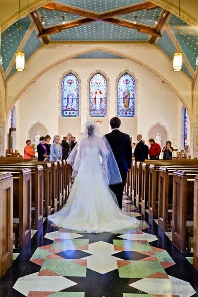 A bride and groom walk down the aisle of a church adorned with stained glass windows. The bride wears a long veil and white dress, while the groom is in a dark suit. Guests are seated in the pews, watching the couple.