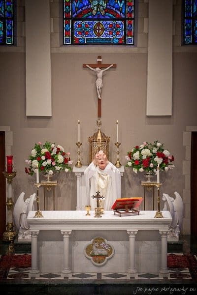 A priest stands at an ornate altar in a church, raising his hands in prayer. The altar is decorated with floral arrangements and candles. Stained glass windows and a crucifix are visible in the background.