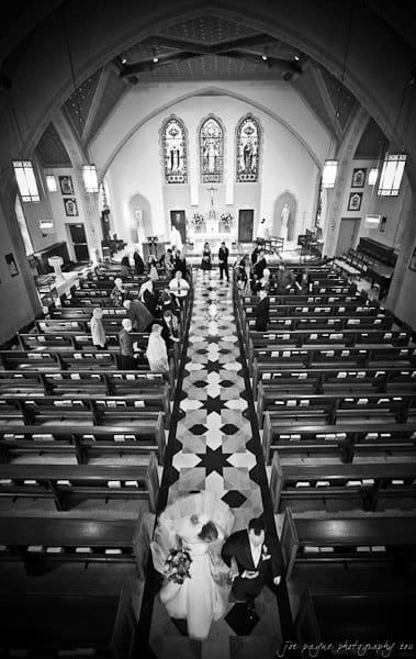 A black-and-white photo of a church interior with a central aisle, stained glass windows, and a checkered floor. A bride and groom walk down the aisle as guests, dressed formally, are seated in wooden pews.