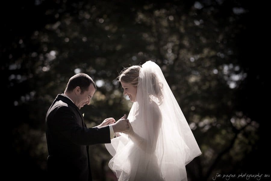 A couple in wedding attire stands smiling and facing each other outdoors. The groom, in a black suit, gently holds the hands of the bride, who is wearing a white gown and veil. Sunlight filters through the surrounding trees, creating a romantic atmosphere.