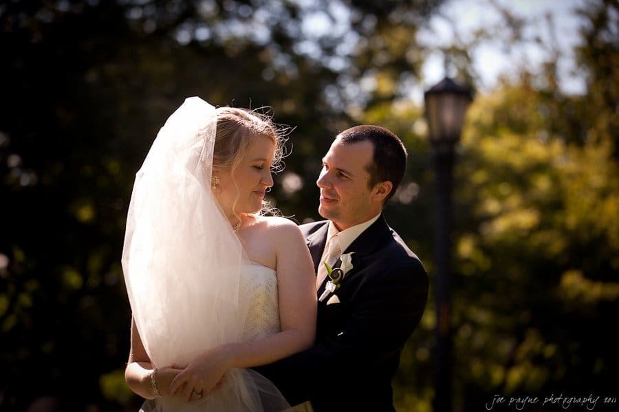 A bride and groom stand outside, embraced closely. The bride wears a white veil and gown, while the groom is in a black suit. They gaze at each other lovingly, surrounded by greenery with a lamppost in the background.