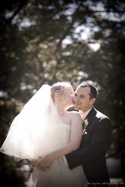A bride and groom embrace outdoors. The bride is wearing a white dress and veil, and the groom is in a dark suit. Sunlight filters through the trees, creating a soft, romantic atmosphere.