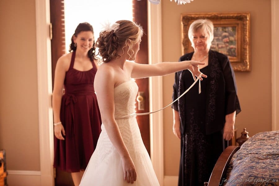 A bride in a white wedding dress holds a ribbon, gesturing playfully. Behind her, a woman in a burgundy dress laughs, and an older woman in a dark outfit smiles warmly. The room is softly lit and decorated with a painting on the wall.
