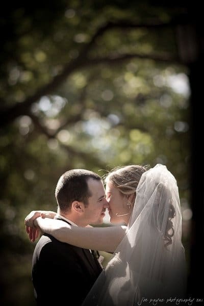 A bride and groom embrace under a canopy of trees. Sunlight filters through the leaves, creating a soft, romantic atmosphere. The bride’s veil cascades elegantly, and both have serene expressions.