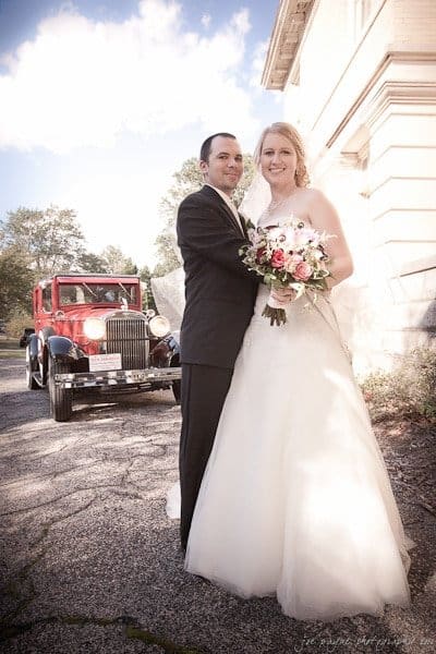 A bride and groom stand happily in front of a vintage red car on a sunny day. The bride holds a bouquet of flowers, and both are elegantly dressed. A large building is in the background.