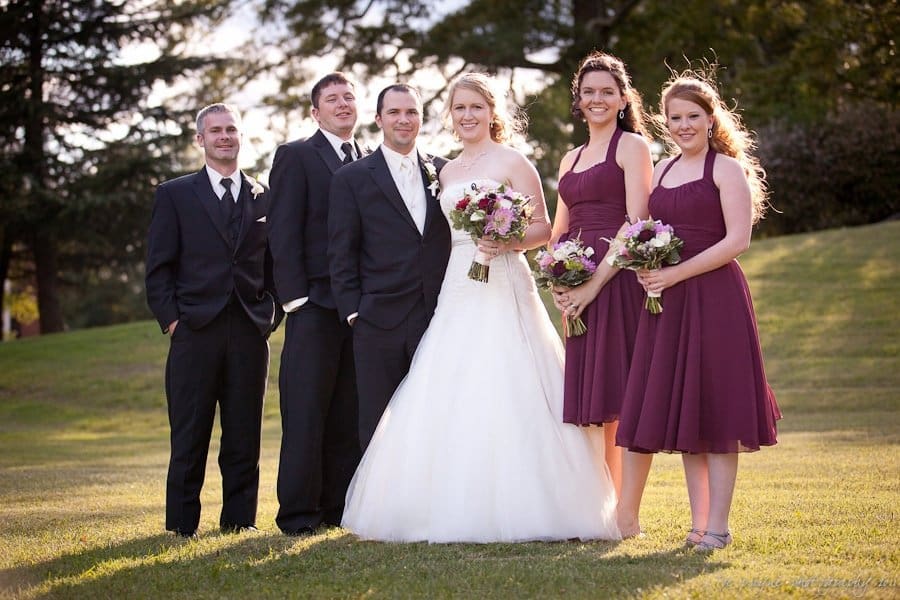 A wedding party posing outdoors on a sunny day. The bride is in a white gown, and the groom is in a black suit. The bridesmaids wear maroon dresses, holding bouquets, while the groomsmen are in black suits. Green grass and trees are in the background.