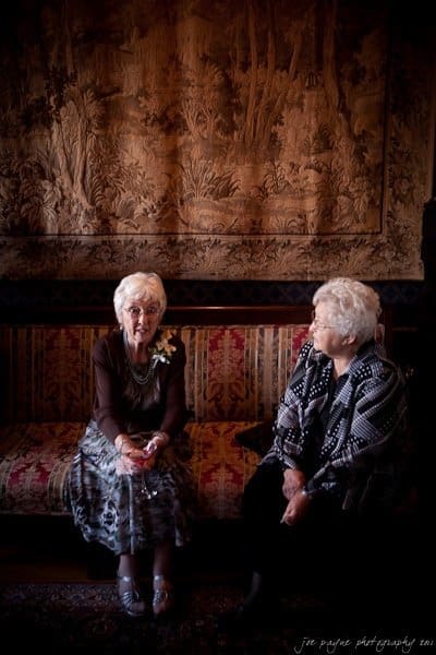 Two elderly women sit on a patterned couch in a dimly lit room with an ornate tapestry hanging in the background. One woman is holding a flower and a drink, while the other is looking at her attentively.