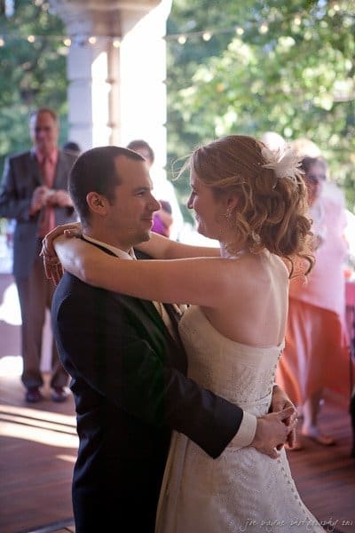 A bride and groom share a dance at an outdoor reception. The bride wears a white gown with her hair in an updo, while the groom is in a dark suit. Guests and greenery are visible in the background.