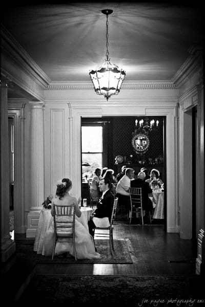 A black and white photo of a bride and groom seated at a small table in a classic interior. Guests are visible in the background, sitting at a larger table. A chandelier hangs above, casting light through the elegant room.