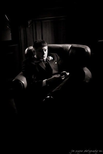 A black and white photo of a person in a suit sitting in an armchair. They appear deep in thought, with a gentle light illuminating their face and hands. The background is dimly lit, creating a dramatic and contemplative atmosphere.