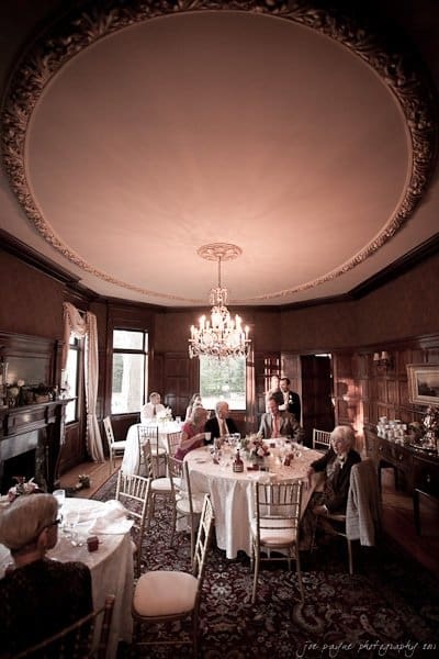 A vintage dining room with ornate chandelier and ceiling molding. Several elegantly dressed people sit around tables covered with white tablecloths, engaged in conversation. The room has dark wooden paneling and a patterned rug.