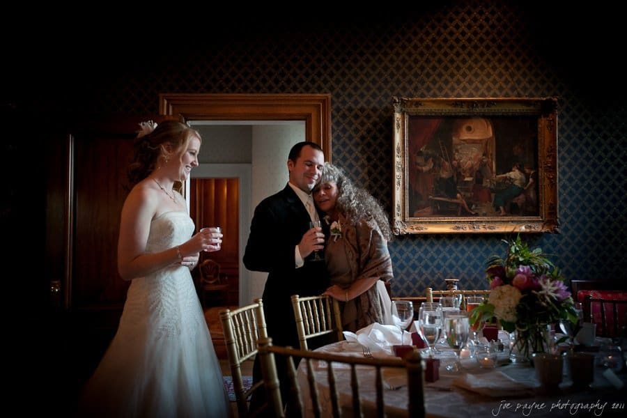 A bride in a white gown stands smiling to the left. A groom in a suit is in the center, embracing an older woman. They are in a dimly lit room with a painting on the wall, and a table with flowers and glassware in the foreground.