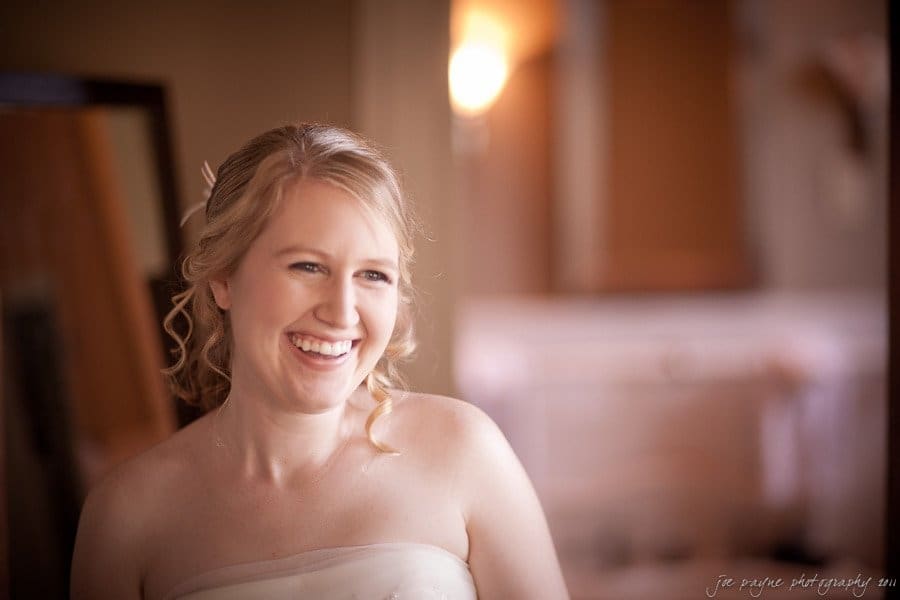 A woman with light hair and a strapless dress smiles warmly while standing indoors. Soft lighting enhances the warm and welcoming atmosphere of the scene.