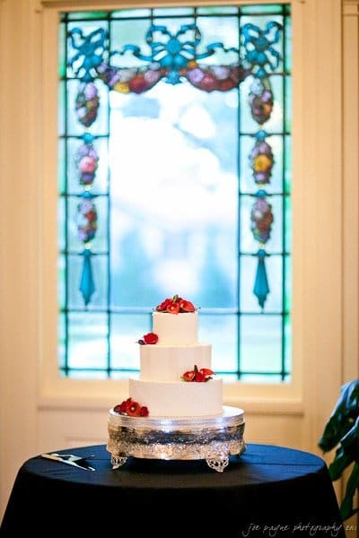 Three-tiered white wedding cake adorned with red flowers sits on a silver stand, placed on a black tablecloth. The background features a stained glass window with colorful floral designs, creating a soft, elegant ambiance.