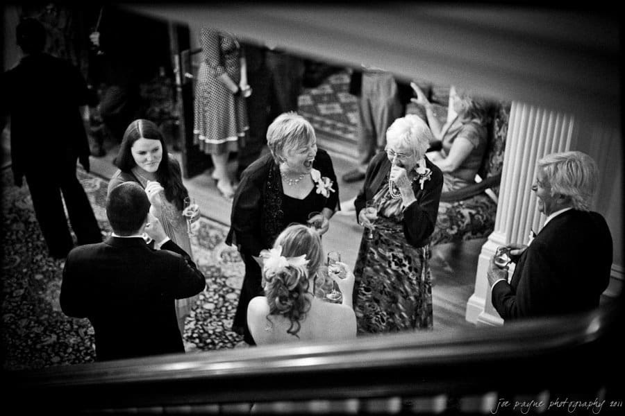 A black-and-white photo shows a group of people at a social event. They are standing in a circle, dressed in formal attire, and engaging in conversation while holding drinks. The setting appears to be an elegant room with patterned carpeting.