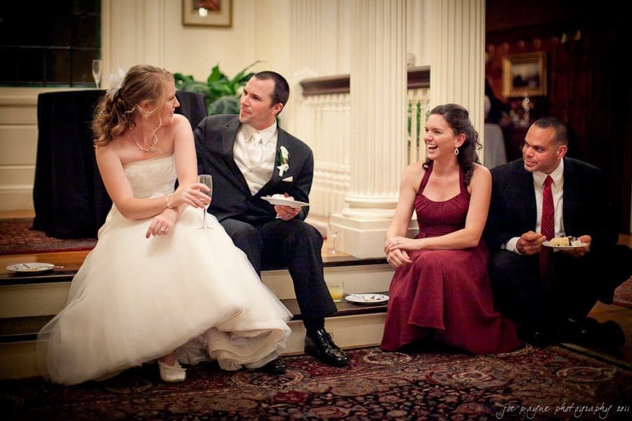 A bride and groom sit on stairs in a warmly-lit room, talking and laughing with two guests. The bride is in a white gown holding a drink, while a female guest in a burgundy dress and a male guest in a suit are holding plates.