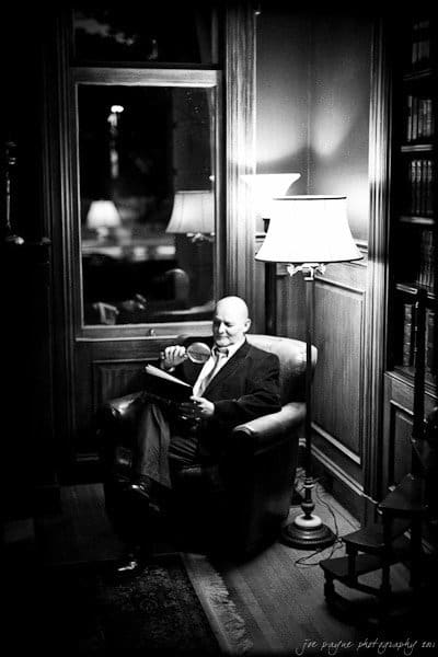A person sits in a leather armchair in a dimly lit room, reading a book with a magnifying glass. They are surrounded by wooden paneling and a tall lamp, with a window in the background. The image is in black and white.