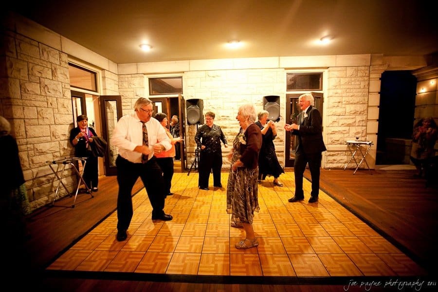 A group of elderly people dance joyfully on a wooden dance floor indoors, with stone walls and windows in the background. The scene is warmly lit, and everyone appears to be enjoying themselves.