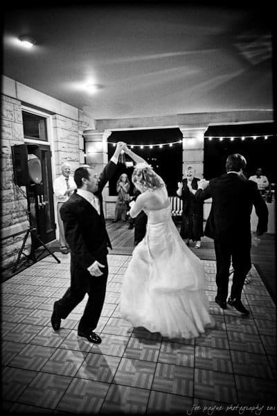 A couple in formal attire dances joyfully on a checkered floor at a wedding reception. The bride wears a flowing white gown, and the groom is in a suit. Guests watch and celebrate in the background under string lights. The setting has a rustic elegance.