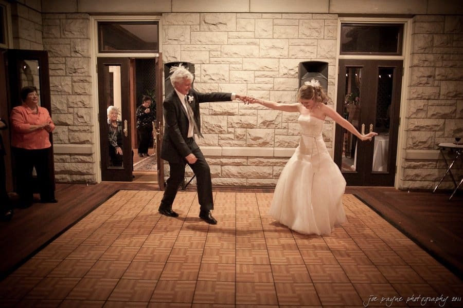 A bride in a white gown and a groom in a dark suit joyfully dance on a wooden floor, surrounded by stone walls. Onlookers stand nearby, watching the celebration.