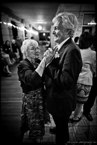 An elderly couple dances closely in a warmly lit room. The woman smiles up at the man as they hold hands. Other people sit and stand in the background, creating a festive atmosphere.