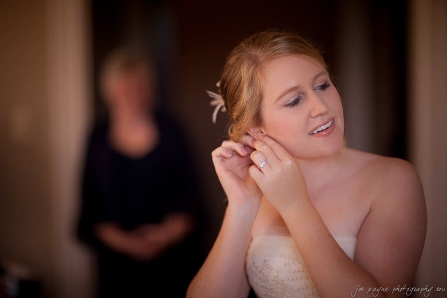 A bride with light brown hair adjusts her earring, smiling softly. She wears a strapless white dress and a delicate hairpiece. A blurred figure stands in the background. The room is warmly lit.