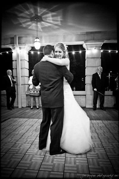 A couple is seen sharing a slow dance on a tiled floor, surrounded by onlookers. The bride is in a white gown and the groom in a dark suit. String lights and columns in the background create a romantic atmosphere. The photo is in black and white.