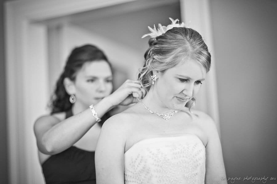 A black and white photo of a bride looking down, smiling slightly, as a bridesmaid helps adjust her hair. The bride wears a strapless dress and a necklace, while the bridesmaid stands focused in the background.