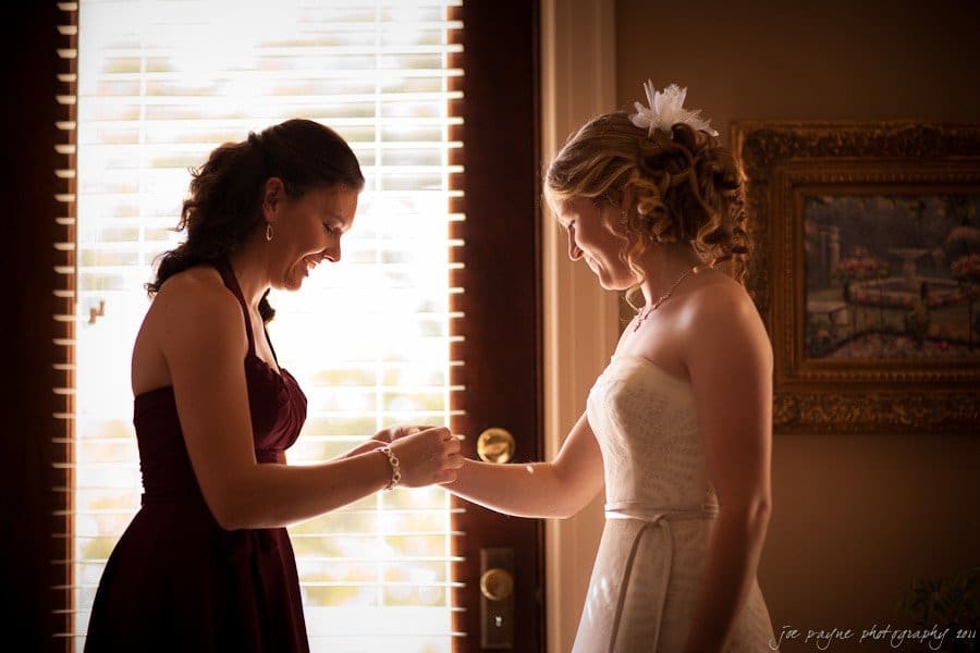Two women stand in front of a window with blinds. The woman on the left wears a dark dress and holds the hand of the woman on the right, who is in a white dress with floral hair accessories. They appear to be smiling and sharing a moment together.