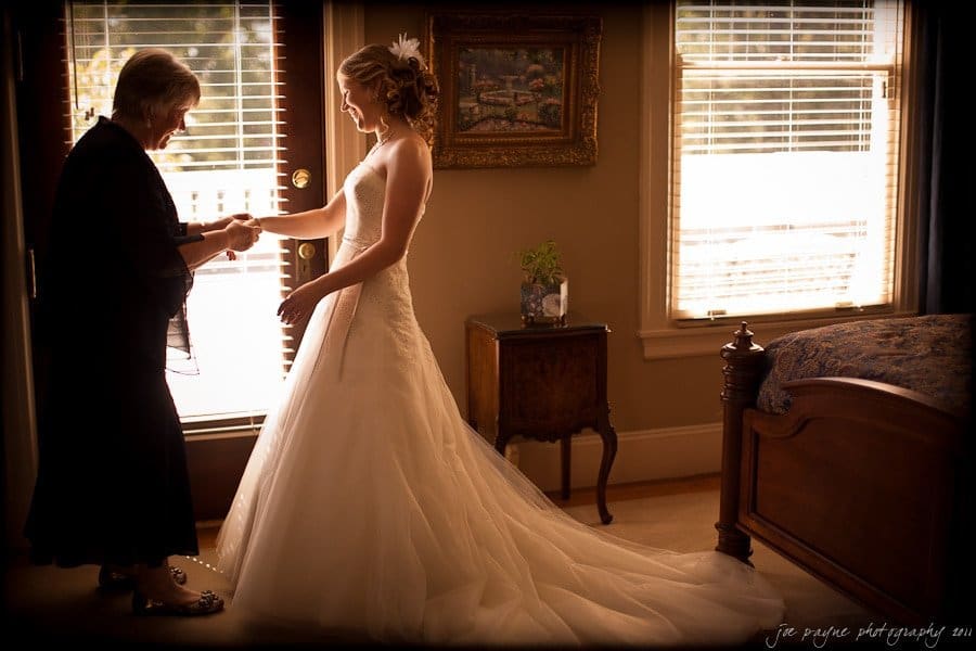 A bride in a white dress stands by a window, holding hands with an older woman in a dark outfit. Sunlight streams in, highlighting the brides dress and creating a warm atmosphere in the elegantly decorated room.