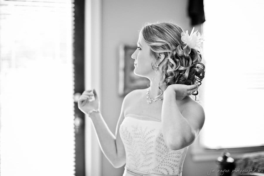 A woman in a strapless dress, with curled hair adorned with a flower, stands indoors. She looks to the side while adjusting her hair with one hand. Natural light from a window softly illuminates her.