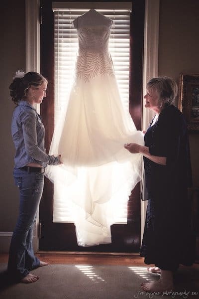 A young woman and an older woman admire a wedding dress hanging in front of a sunlit window. The young woman, dressed casually, stands on the left, while the older woman, in a dark outfit, stands on the right.