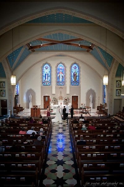 A wedding ceremony inside a church with stained glass windows. The bride and groom stand at the altar, accompanied by the officiant and a few guests seated in the pews. The ceiling is adorned with wooden beams and soft lighting.