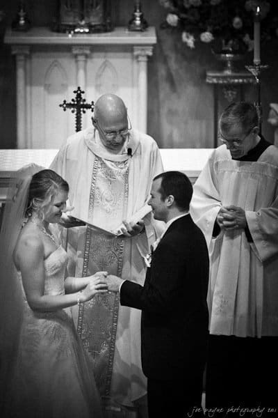 A bride and groom exchange rings in a church ceremony. The bride is in a white gown, and the groom is in a black suit. Two clergymen officiate, one holding a book. A cross is visible in the background. The scene is in black and white.