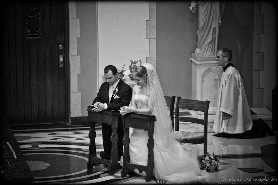 A bride and groom kneel at an altar during a wedding ceremony, with a priest seated nearby in a church setting. The bride wears a traditional gown and veil, while the groom is in a suit. Floral arrangements are visible on the floor.
