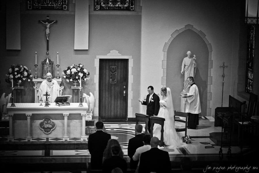A black and white photo of a church wedding ceremony. The couple stands before a priest at the altar, with another man beside them. Guests are seated in the foreground. The altar is decorated with flowers and religious symbols.